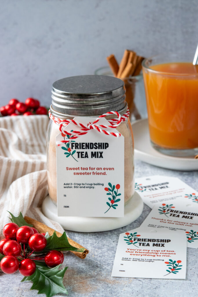 A jar of homemade friendship tea mix decorated with Christmas ribbon and a Christmas gift tag. In the back is a cup of the hot orange spiced tea in a mug.