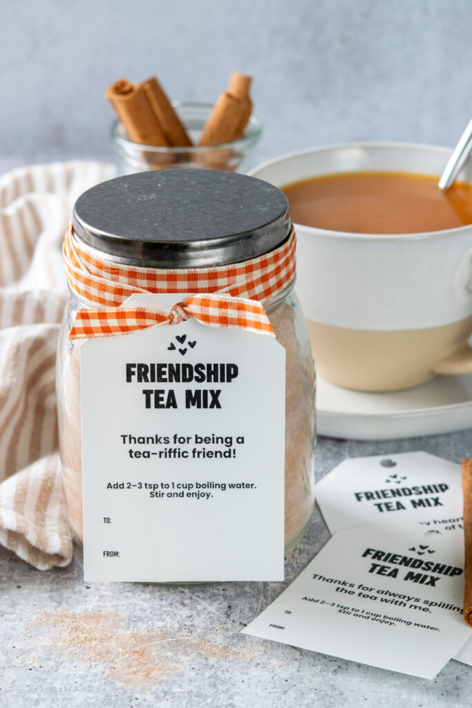 A jar of homemade friendship tea mix decorated with orange ribbon and a gift tag. In the back is a cup of the orange spiced tea in a tea cup.