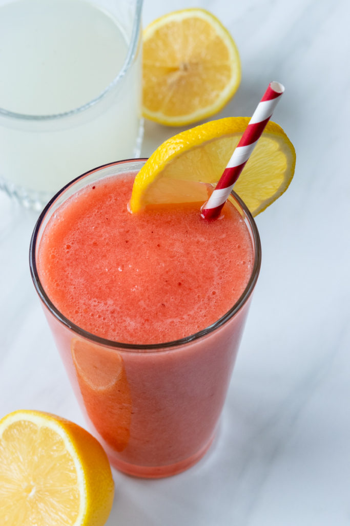 homemade blended strawberry lemonade in tall glass with sliced lemons and cup of lemonade