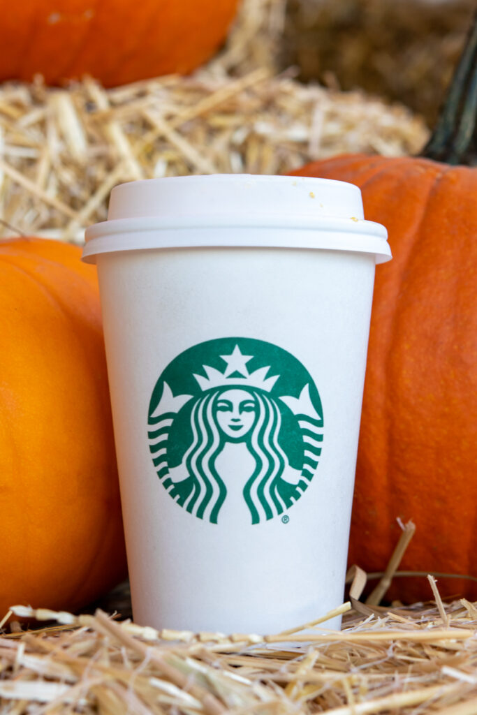 A Starbucks pumpkin spice milk steamer in a Starbucks cup with pumpkins behind the drink.