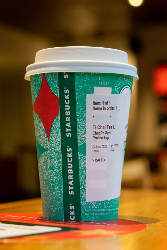 A Starbucks holiday cup showing the order sticker for a tall chai tea latte with chestnut praline syrup and praline topping added in.