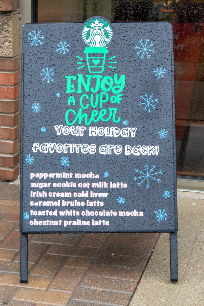 A Starbucks hand-drawn sign that says enjoy a cup of cheer and has a list of favorite holiday drinks.