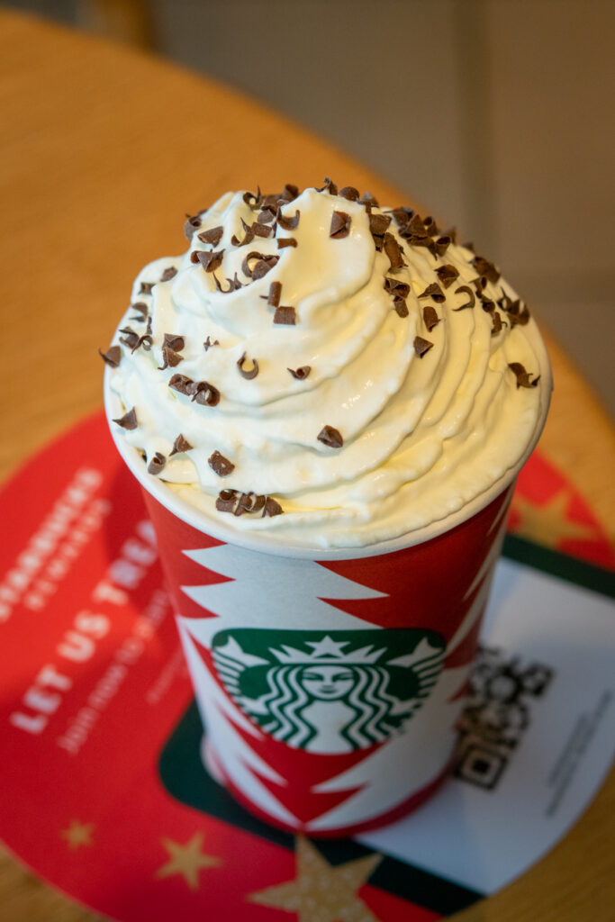 Starbucks hot chocolate in a holiday cup with lots of whipped cream and chocolate curls on top.