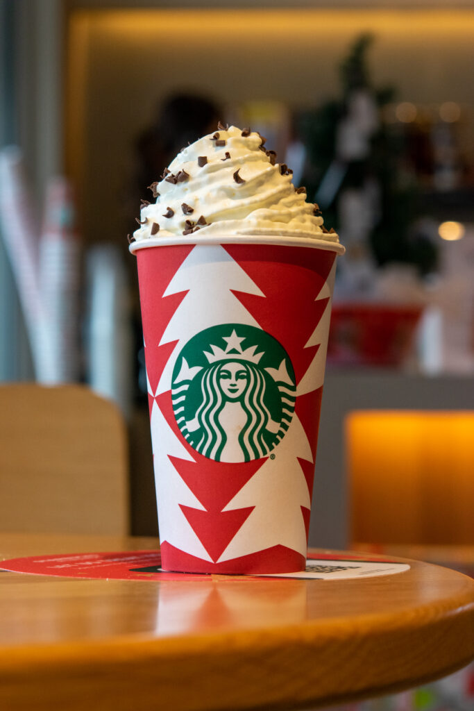 A hot chocolate in a grande Starbucks red cup with a big dollop of whipped cream on top.