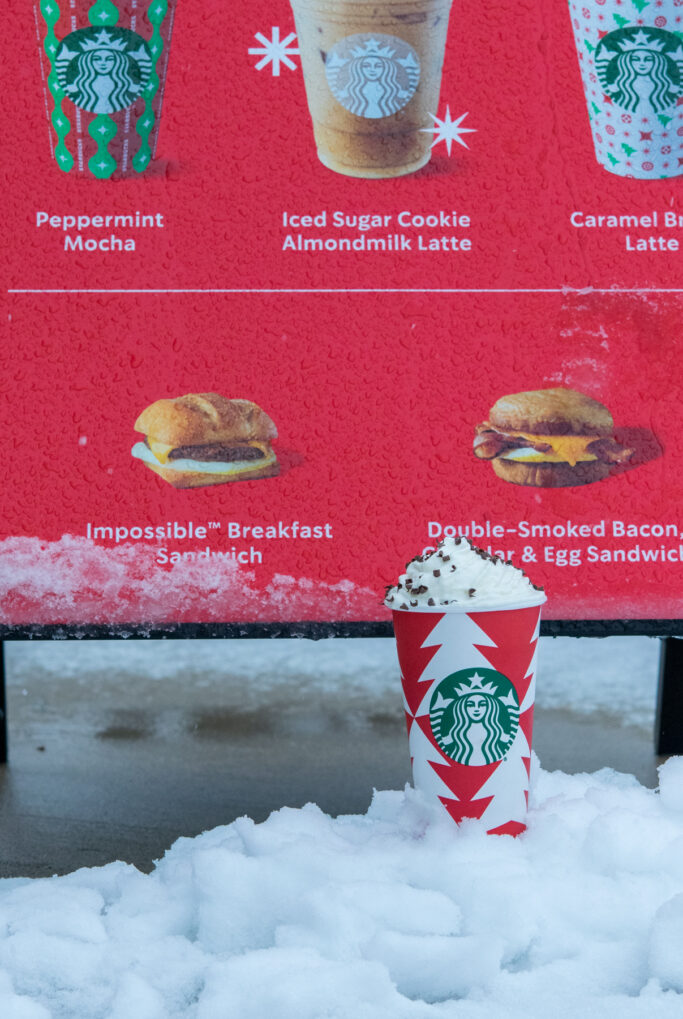 Cup of Starbucks hot chocolate sitting in the snow in front of a Starbucks holiday menu board.
