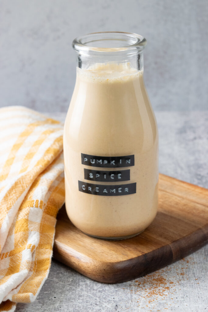 A small glass bottle filled with homemade pumpkin spice creamer. The bottle has a label on it that says, pumpkin spice creamer, and it is sitting on a wood board with an orange checked towel next to it.