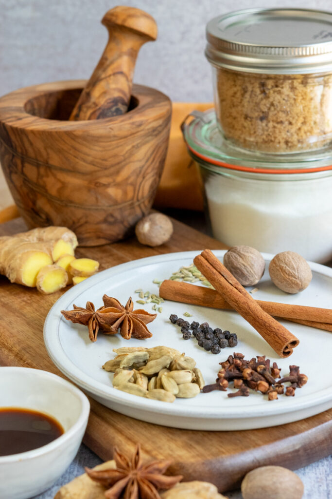 All the whole spices needed to make chai syrup, sliced ginger root, jars of granulated and brown sugars, and a bowl of vanilla syrup.