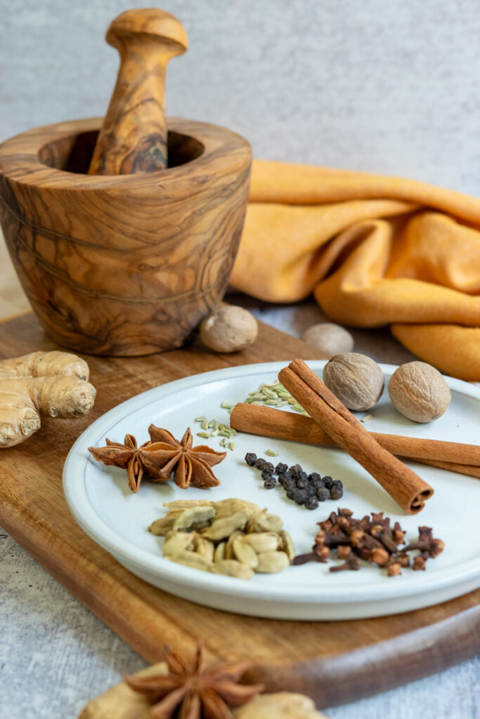 Whole spices, including cinnamon sticks, nutmeg, star anise, cardamom, cloves, black peppercorns and ginger, sitting on a plate and cutting board.