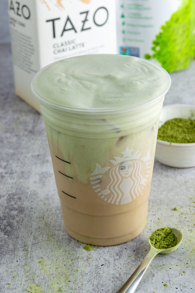 A copycat Starbucks Iced Chai Tea Latte with matcha cold foam in a Starbucks cup, near a box of Tazo chai tea concentrate and a bowl of matcha.