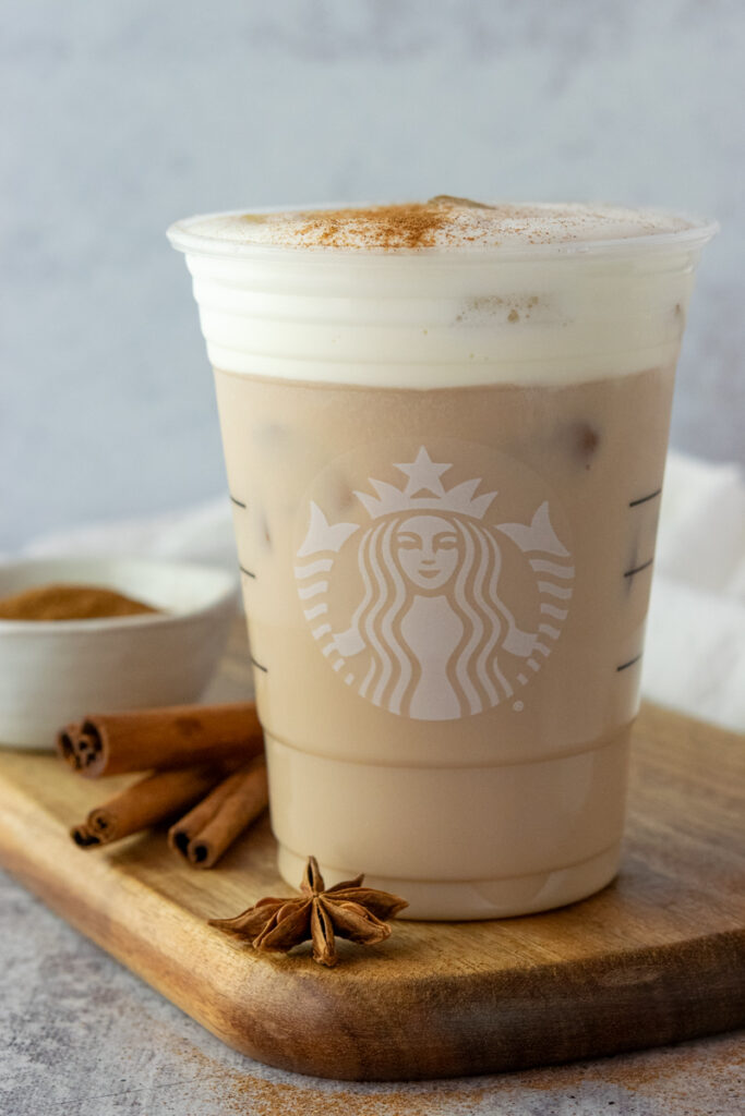 An iced chai tea latte with vanilla sweet cream cold foam made at home in a Starbucks cup with a bowl of cinnamon and cinnamon sticks in the background.