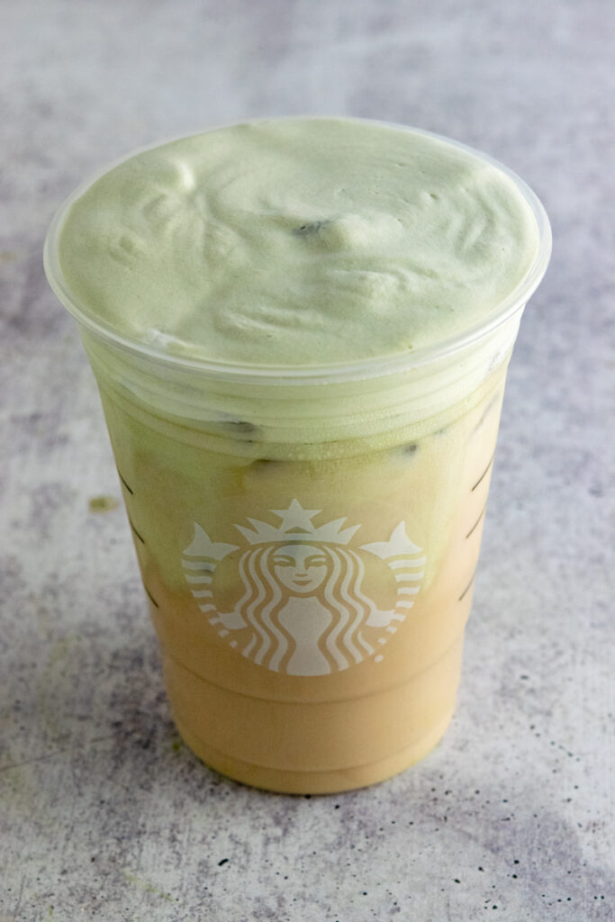 A creamy layer of thick matcha cold foam on top of an iced chai tea latte made at home in a Starbucks grande cup.
