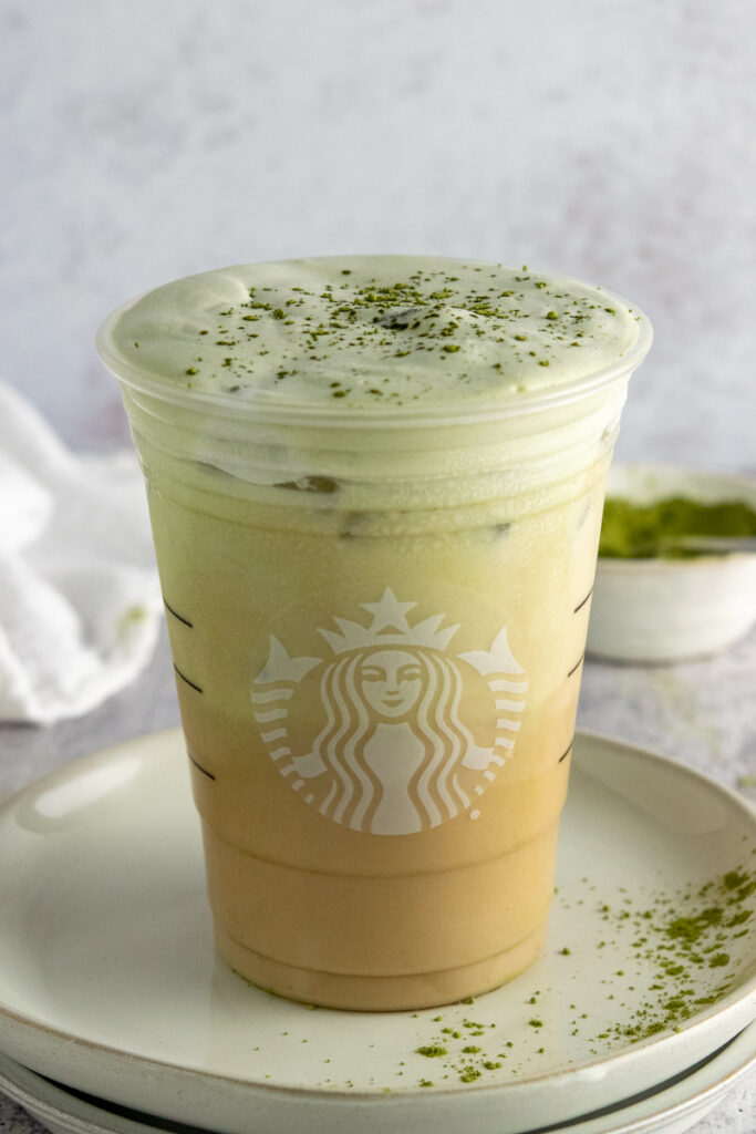 Matcha cold foam layered on top of an iced chai tea latte with a little matcha sprinkled on top in a Starbucks cup.