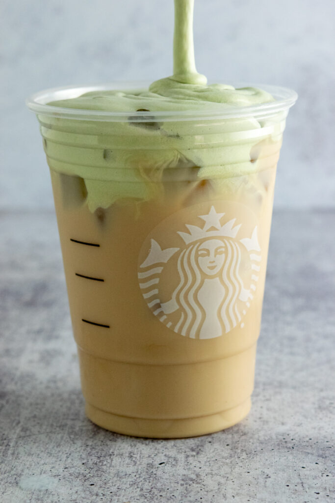 Thick matcha cold foam being poured on top of an iced chai tea latte in a Starbucks cup.