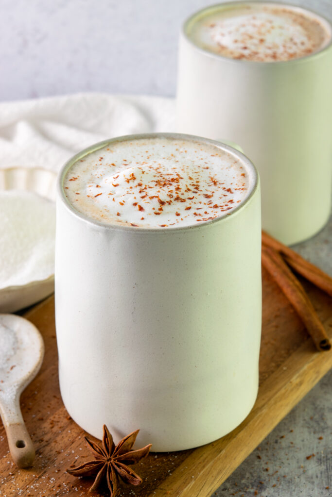 Two chai tea lattes in mugs next to a bowl of sugar and cinnamon sticks.