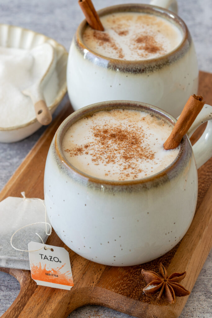 A chai tea latte in a mug with cinnamon sprinkled on top  and a cinnamon stick in it, sitting next to a chai tea bag and whole spices.