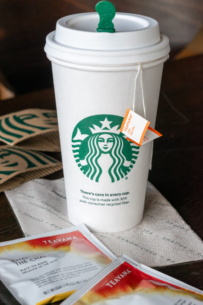 A grande Starbucks cup with two chai tea bags hanging out of it, sitting on a table in Starbucks near two Teavana chai tea bag wrappers and a cup sleeve for hot drinks.