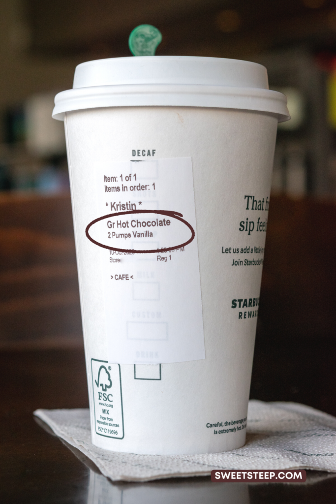 A hot chocolate ordered from Starbucks showing the order sticker on the cup with 2 pumps of vanilla syrup added to the drink.