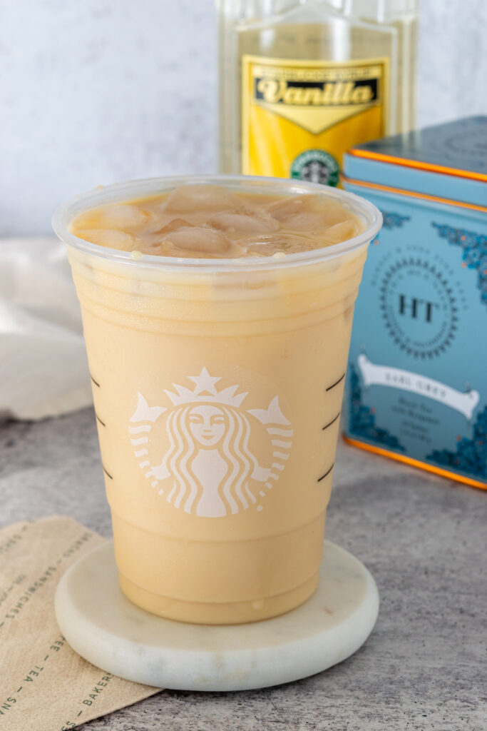 A homemade iced London Fog in a Starbucks cup sitting in front of a tin of earl grey tea bags and bottle of Starbucks vanilla syrup.