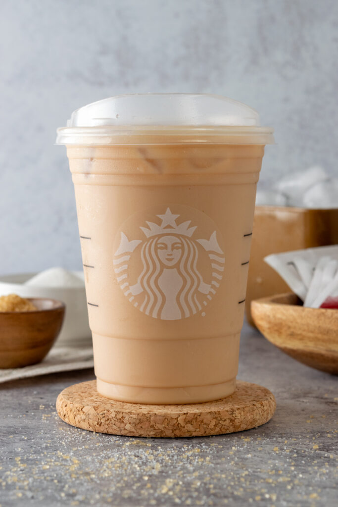 An ice cold breakfast tea latte made at home, in a Starbucks cup inf front of sugar bowls, and scoop of ice cubes.