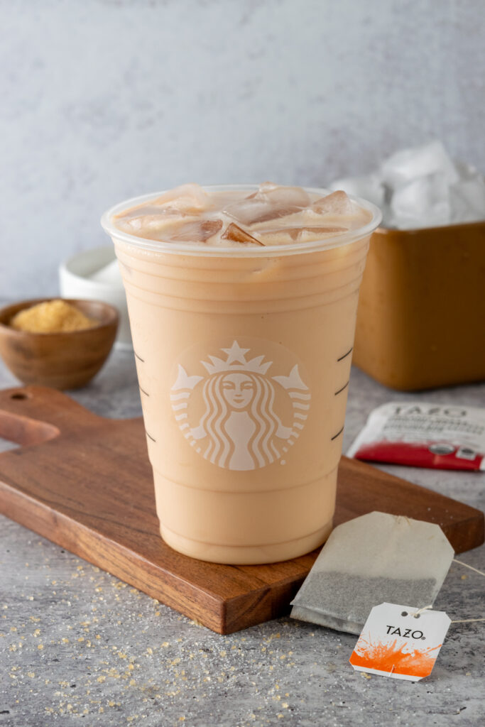 A homemade iced English Breakfast tea latte in a Starbucks cup in front of bowls of sugar, a big scoop of ice cubes and two tea bags.