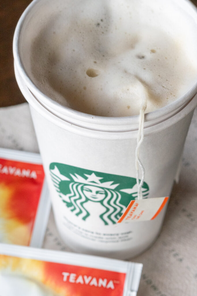 A Starbucks cup showing a frothy chai tea latte with chai tea bags in it.