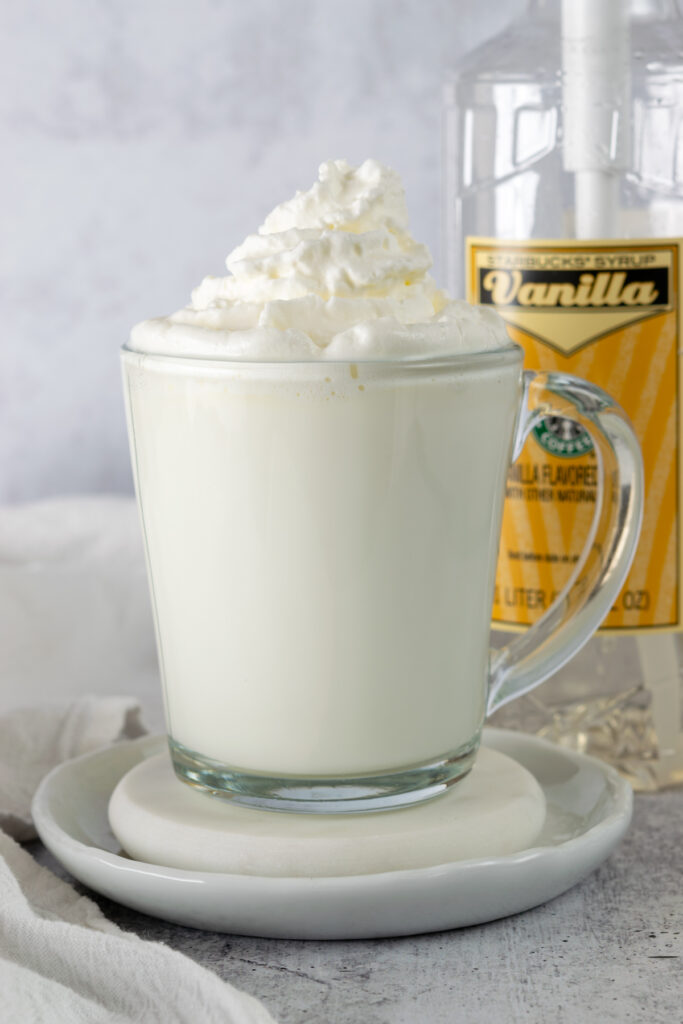 A hot vanilla steamer in a glass mug in front of a bottle of Starbucks vanilla syrup.