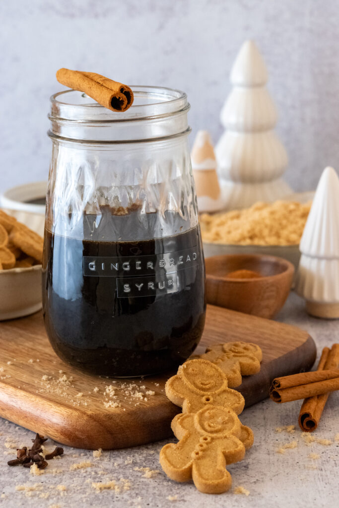 Jar of homemade gingerbread syrup with cinnamon stick on top and gingerbread cookies, cinnamon sticks and cloves next to jar.