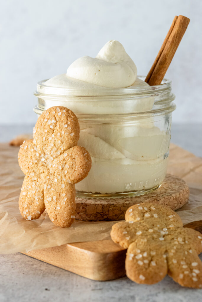 Homemade gingerbread whipped cream swirled in a small glass jar with a cinnamon stick in it and gingerbread cookies in front of it.