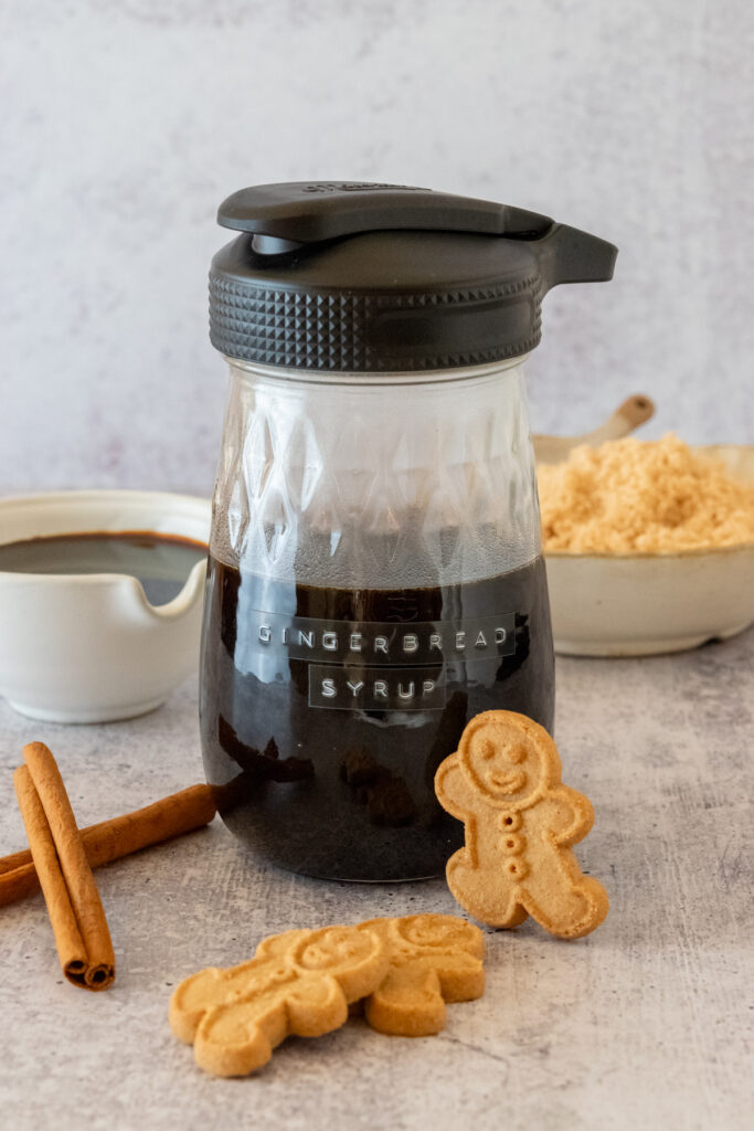 A gingerbread cookie leaning against a bottle of homemade gingerbread syrup which is next to small bowls of molasses and brown sugar.