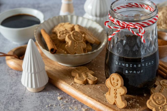 Jar of homemade gingerbread syrup surrounded by gingerbread cookies, bowl of molasses and small decorative Christmas trees.