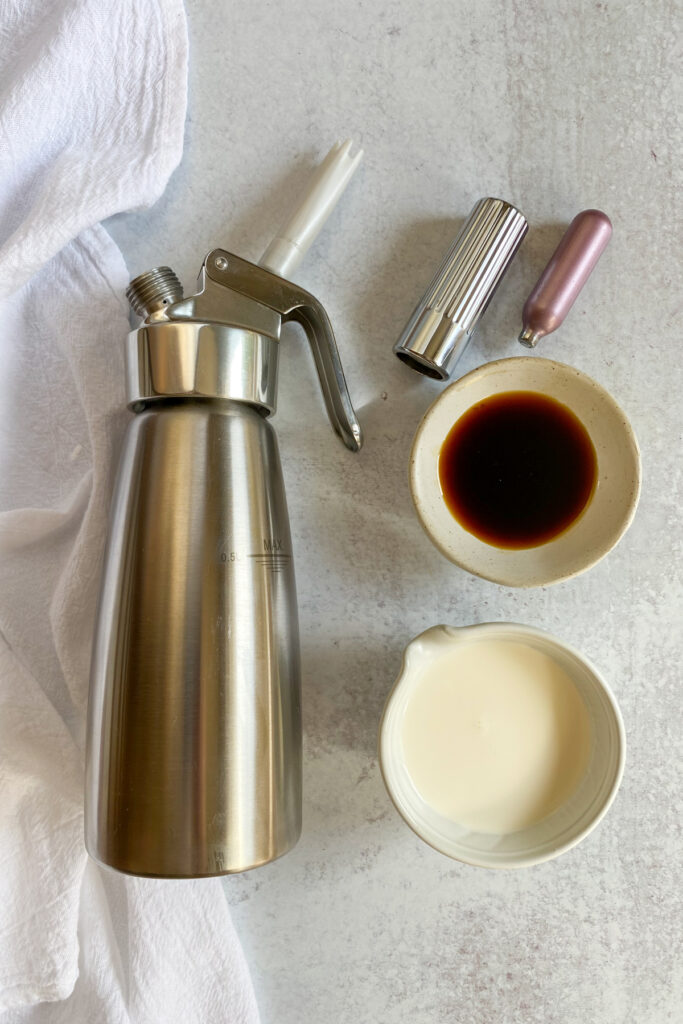 A whipped cream whipper, charger, charger holder and bowls of gingerbread syrup and heavy cream.