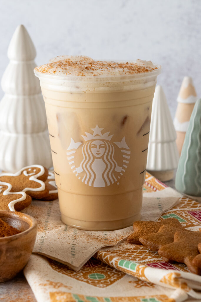 A Starbucks copycat Iced Gingerbread Oat Milk Chai Tea Latte in a Starbucks cup with small decorative Christmas trees in the background and gingerbread cookies surrounding the cup.