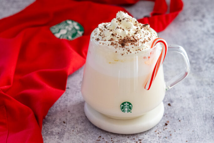 Starbucks copycat peppermint white hot chocolate with a candy cane on rim next to a red holiday Starbucks apron.