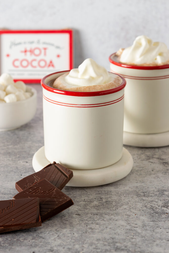 Two mugs of homemade hot chocolate topped with whipped cream and dark chocolate squares leaning against one cup.