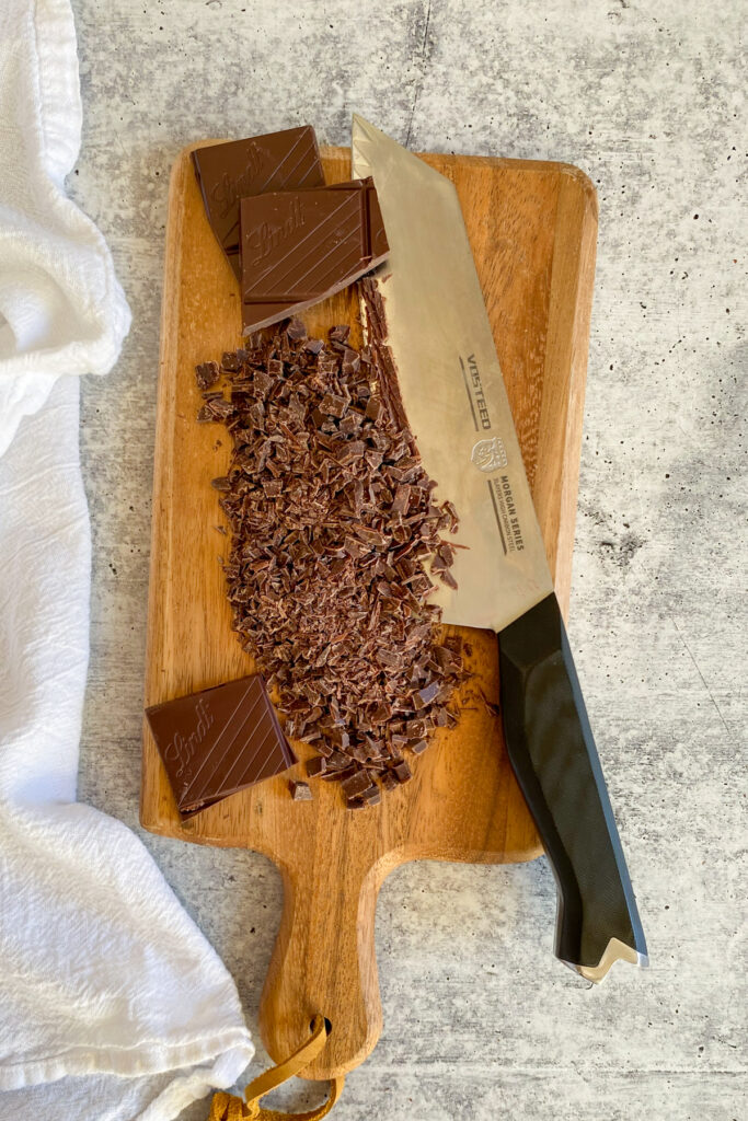 Chopped chocolate bar and a knife on a wooden cutting board.