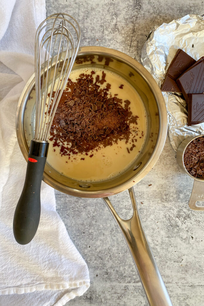 Chopped chocolate added to saucepan with heavy cream with chocolate squares and a whisk next to it.
