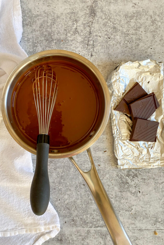Melted chocolate in a saucepan next to a whisk and chocolate squares on a foil wrapper.