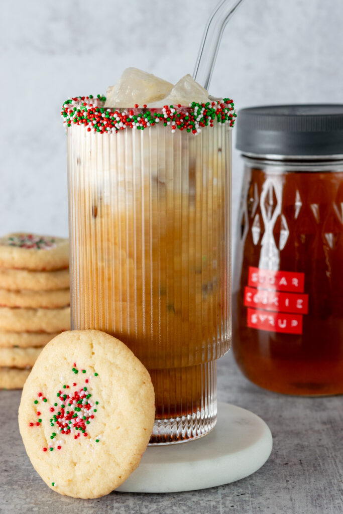 An iced sugar cookie latte served in a festive glass that has sprinkles on the rim. There is a sugar cookie leaning on the glass and the bottle of syrup behind the drink.
