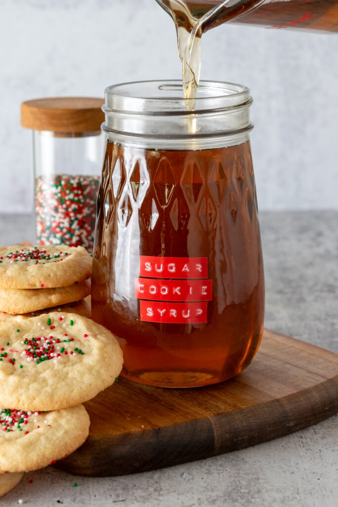 The cookie syrup has been transferred to a glass jar. There are some sugar cookies with sprinkles on top and a small jar of red, green and white sprinkles next to the syrup bottle.