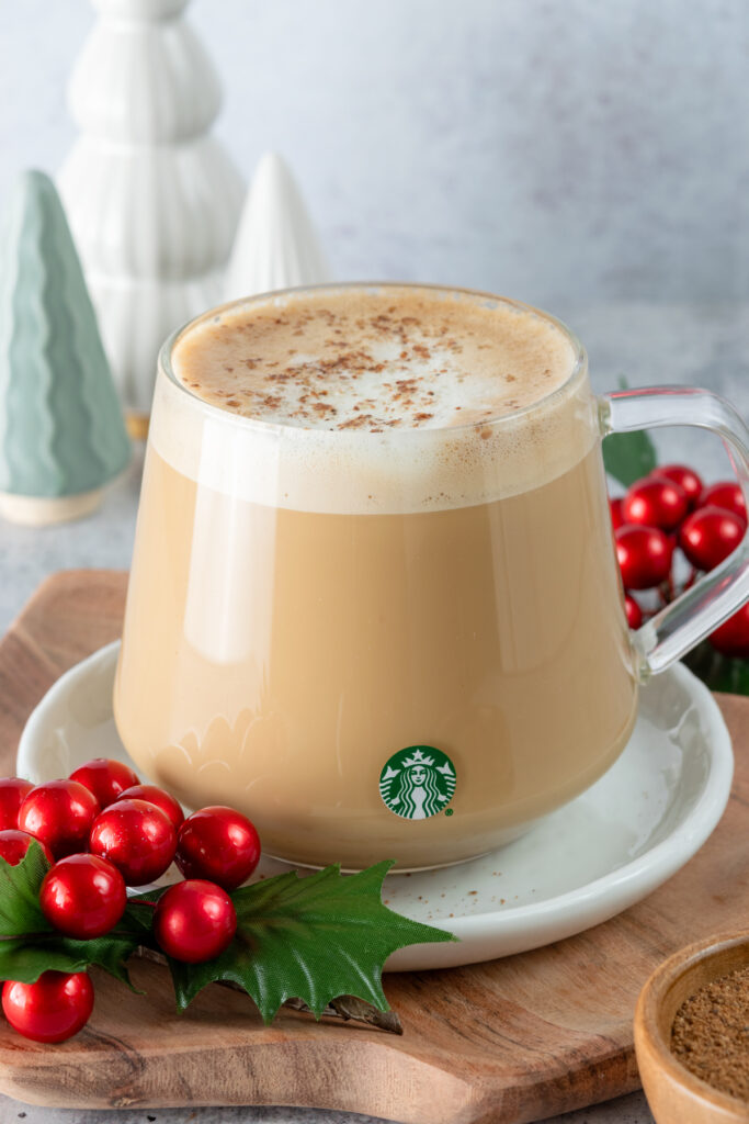 A homemade eggnog latte in a Starbucks cup with a small bowl of nutmeg next to it and Christmas tree decoration in the background.