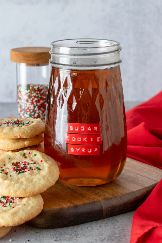 A bottle of sugar cookie coffee syrup made at home. The flavored syrup bottle has a red label on it and is next to a stack of sugar cookies and a bottle of red, green and white sprinkles.