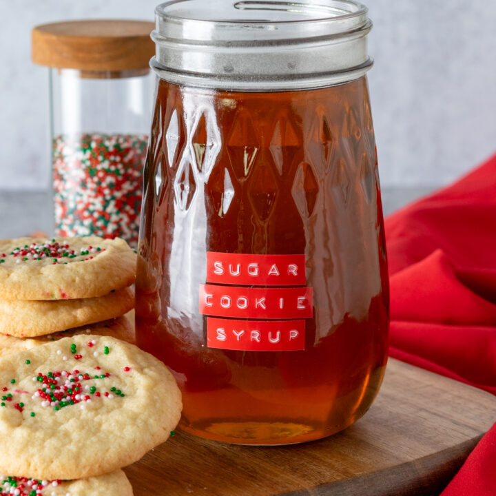 A bottle of sugar cookie coffee syrup made at home. The flavored syrup bottle has a red label on it and is next to a stack of sugar cookies and a bottle of red, green and white sprinkles.