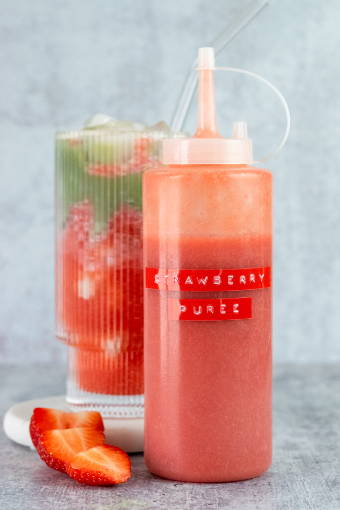Homemade strawberry puree for drinks in a squeeze bottle with label that says strawberry puree, next to an iced matcha latte layered with the puree, milk and matcha.