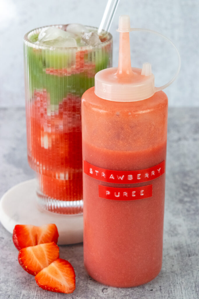 Bottle of homemade strawberry puree next to an iced strawberry matcha latte and a sliced strawberry.