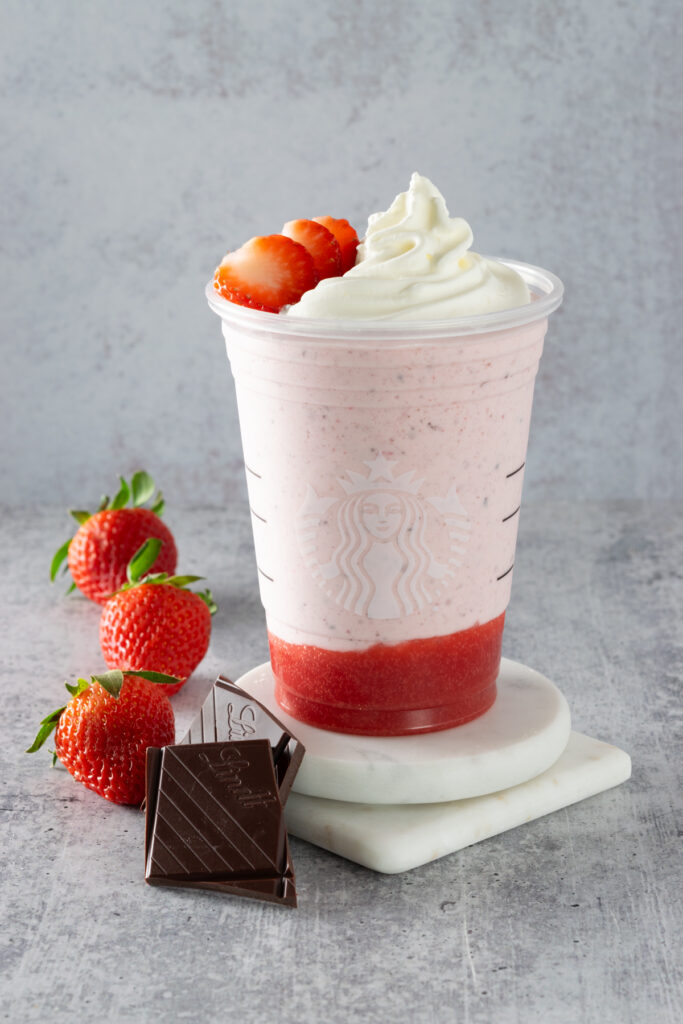 A copycat Starbucks Chocolate-Covered Strawberry Frappuccino in a Starbucks cup sitting next to three fresh strawberries and pieces of dark chocolate.