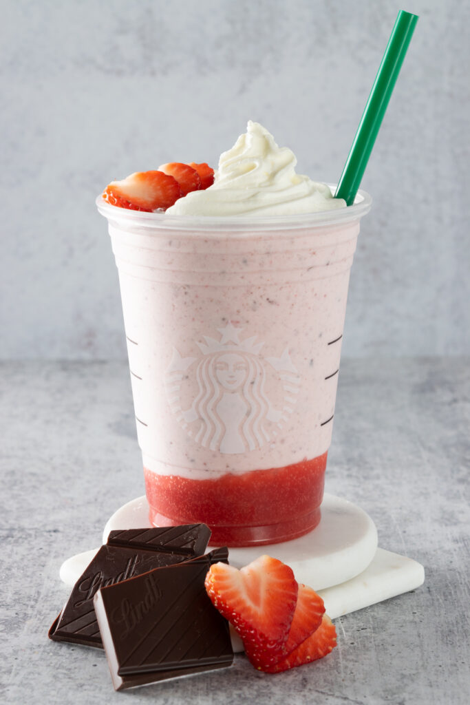 Homemade Starbucks Chocolate-Covered Strawberry Creme Frappuccino with a green straw and a sliced strawberry and chocolate bar sitting next to cup.