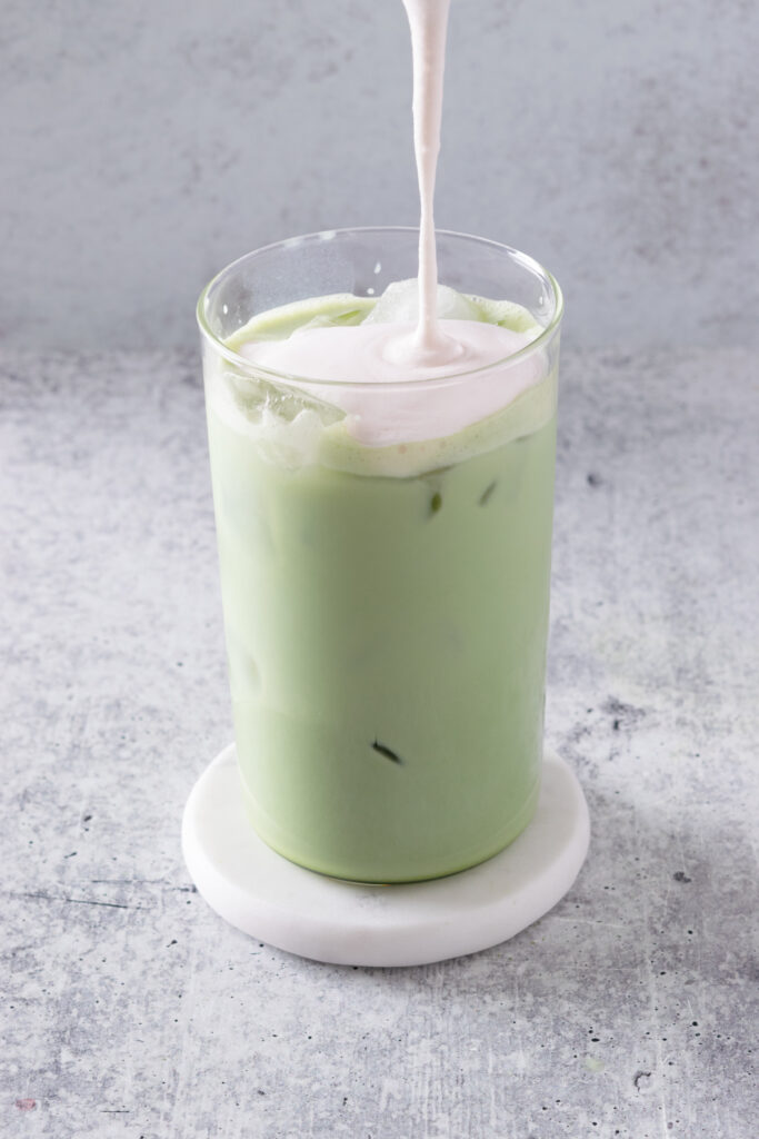 Stream of thick strawberry cream cold foam being poured on top of iced matcha latte.
