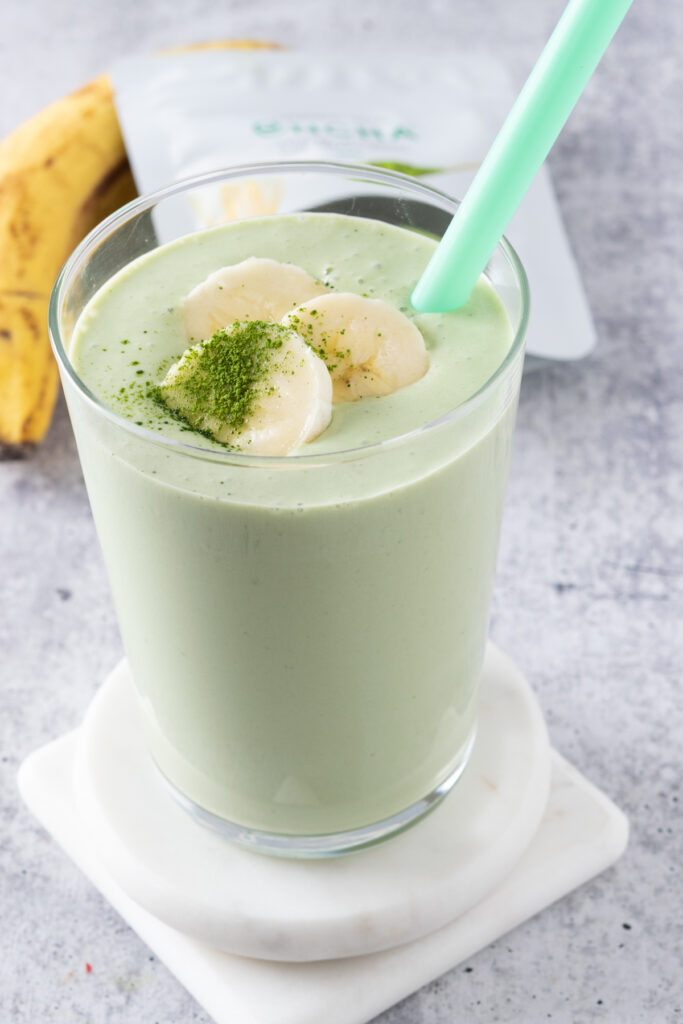 A homemade banana matcha smoothie  garnished with banana slices and a little matcha powder sprinkled on top, in a glass with a green straw sitting in front of a ripe banana and container of matcha green tea powder.