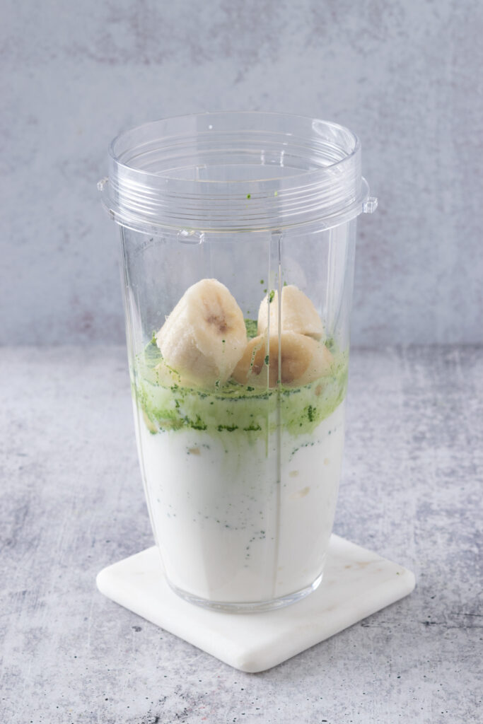 Frozen banana added to blender cup along with matcha, milk and Greek yogurt.