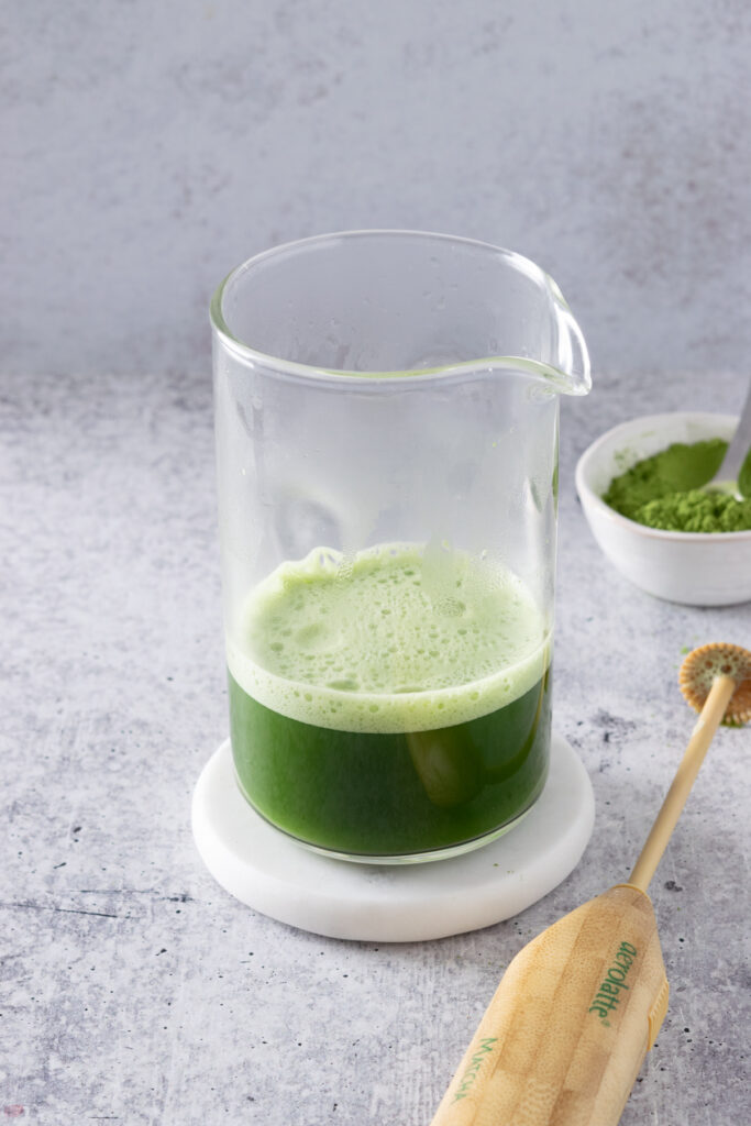 A tall glass with spout that has frothy whisked matcha in it, next to a small bowl of matcha powder and an aerolatte matcha frother.